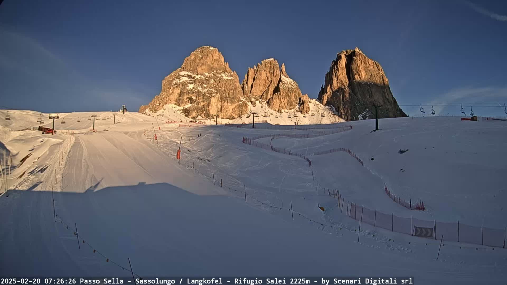 A snow-covered ski slope with chairlifts and rocky mountains in the background under a clear blue sky.