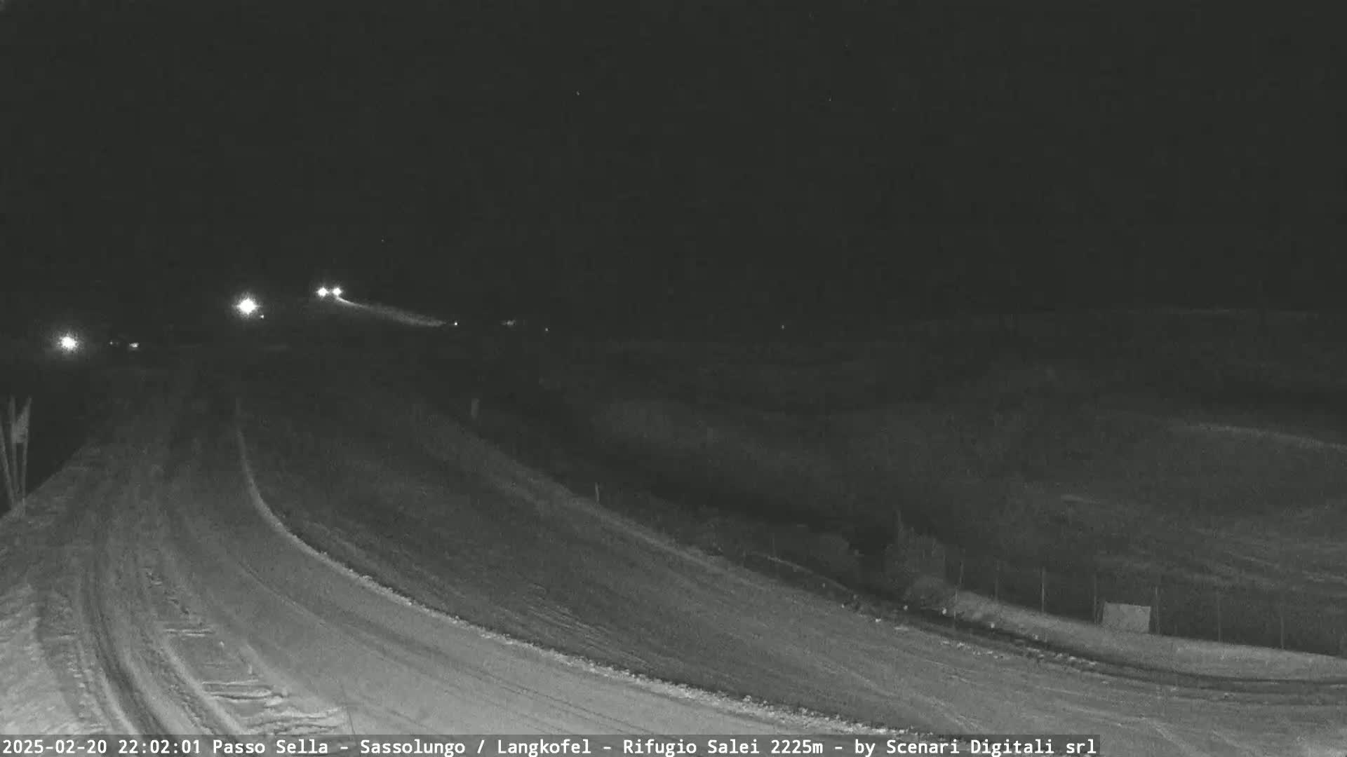 A nighttime, snow-covered road curves gently upward, with a few vehicles visible in the distance under a dark sky.