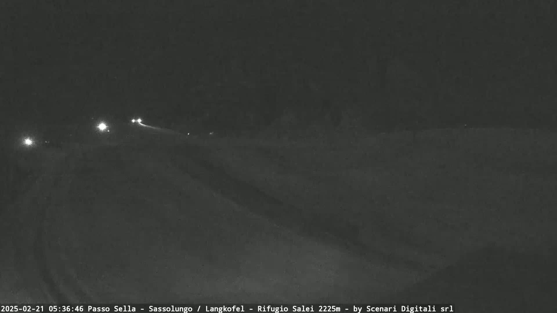 A dark, nighttime view of a snow-covered road with a few vehicles visible in the distance under a clear sky.