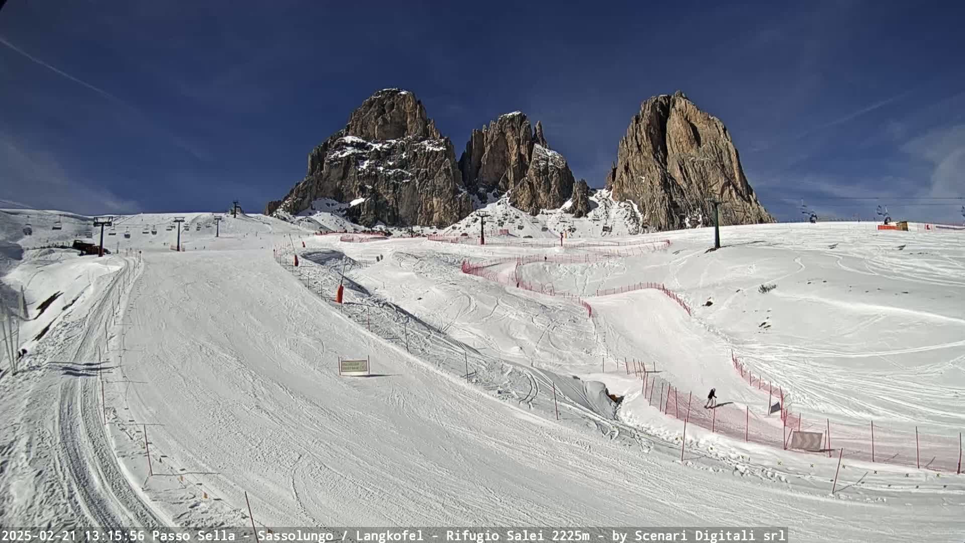 A snow-covered ski slope with several ski lifts and runs, set against a backdrop of towering, snow-dusted rocky mountains under a clear blue sky.