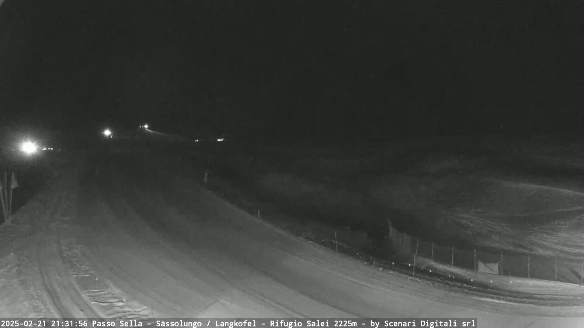 A snow-covered road at night is dimly lit by vehicle headlights, with a slightly hilly landscape visible in the background under a dark sky.
