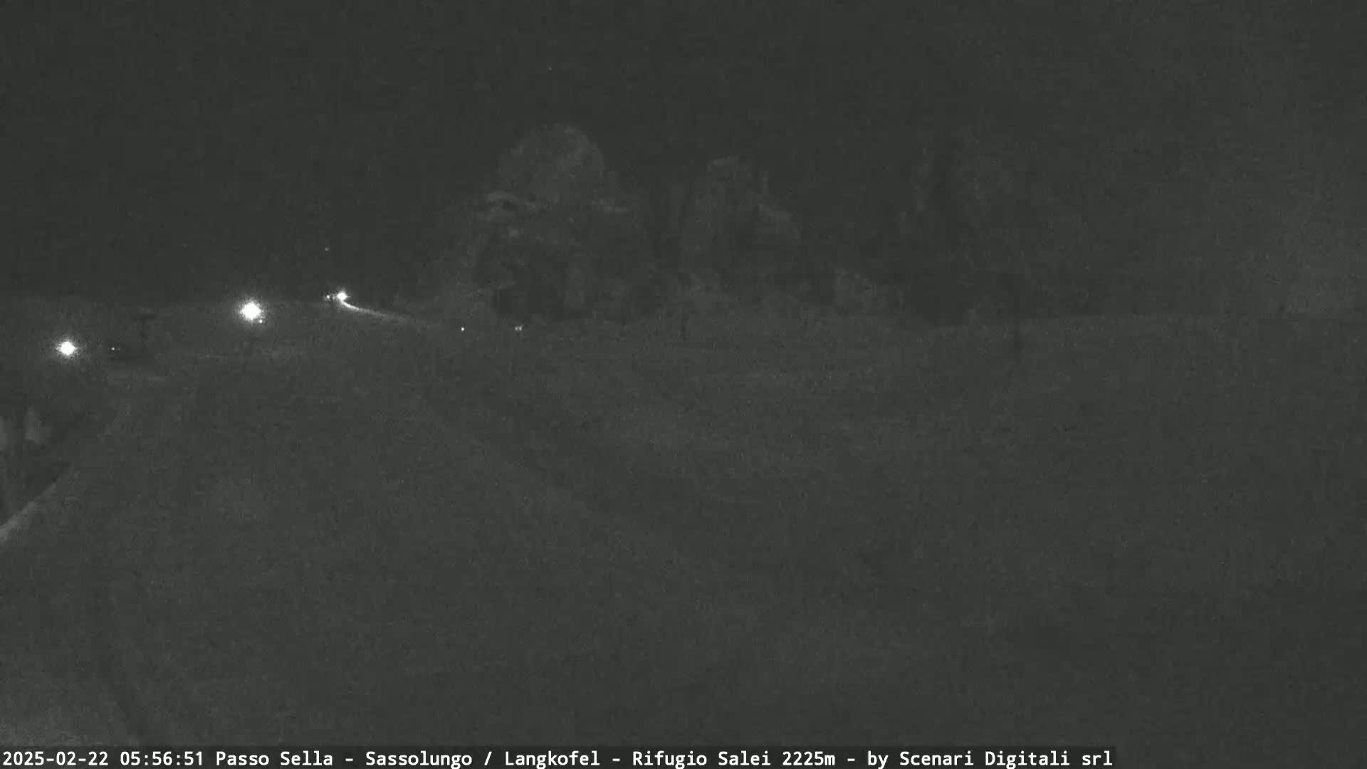 At night, several vehicles with headlights on drive along a road past dark trees and a mostly-empty landscape.