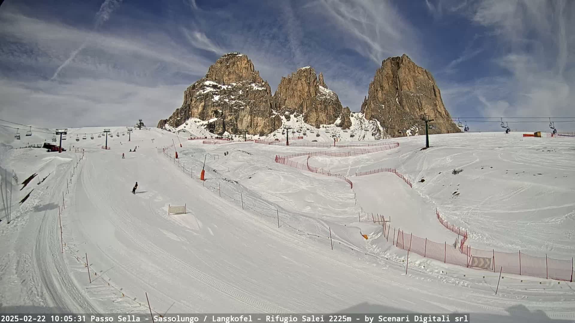A snowy ski slope with several chairlifts and rocky mountain peaks in the background under a partly cloudy sky.