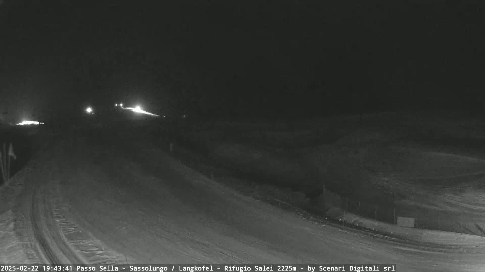A dark, snow-covered road at night is illuminated by the headlights of a few distant vehicles.