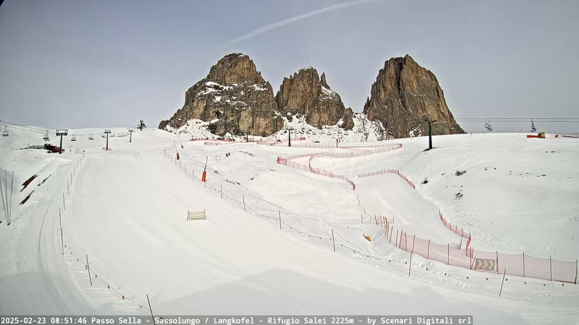 A snow-covered ski slope with several ski lifts and rock formations in the background under a mostly clear sky.