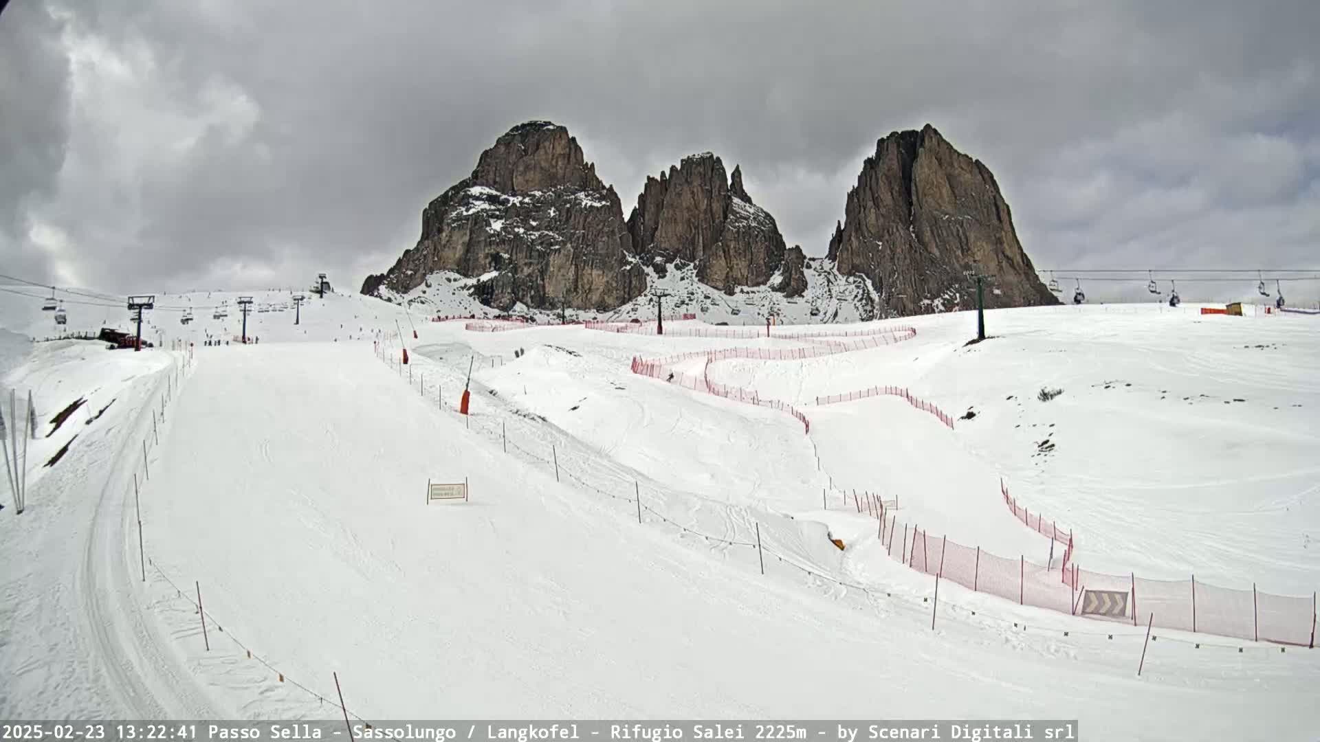 A snow-covered ski slope with chairlifts and red safety nets is nestled beneath a cloudy sky and imposing, rocky mountain peaks.