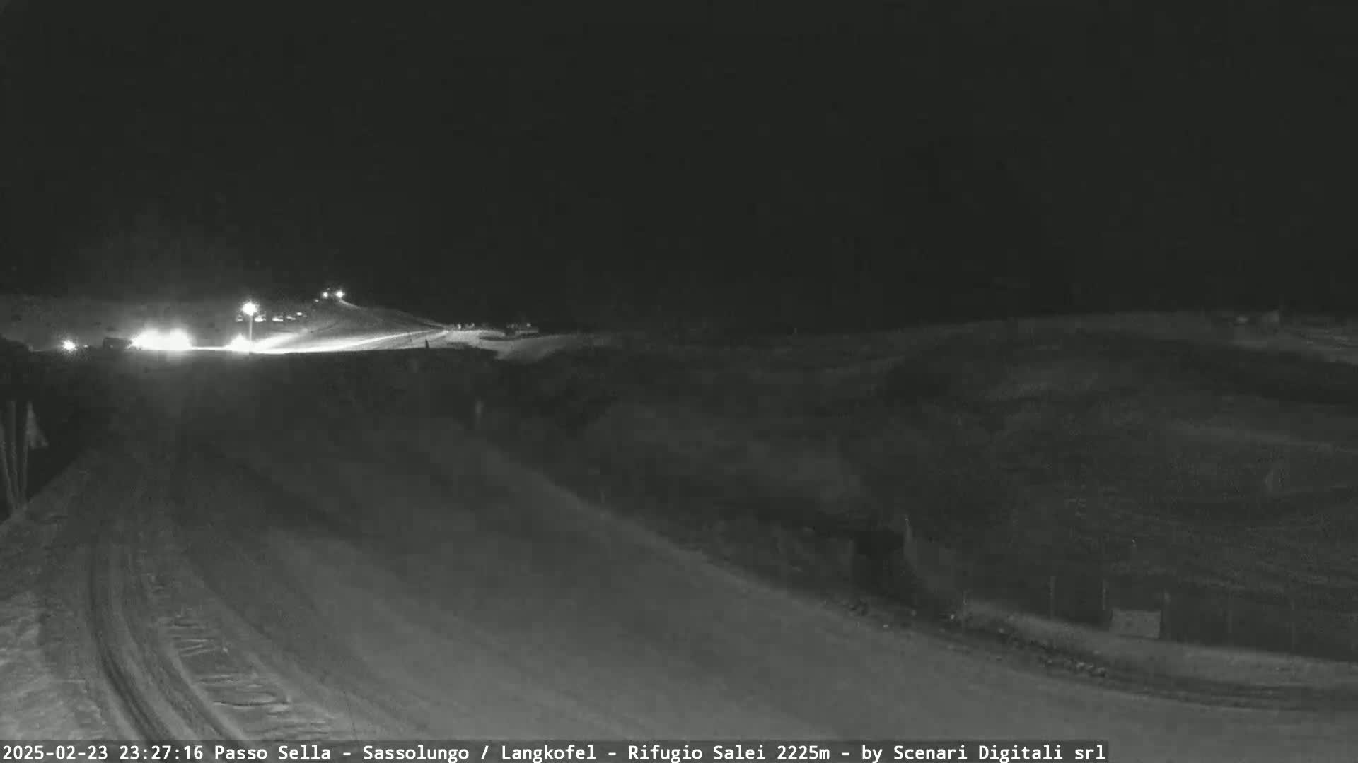 A nighttime, snow-covered road scene shows several vehicles with headlights on driving uphill toward a dimly lit area.