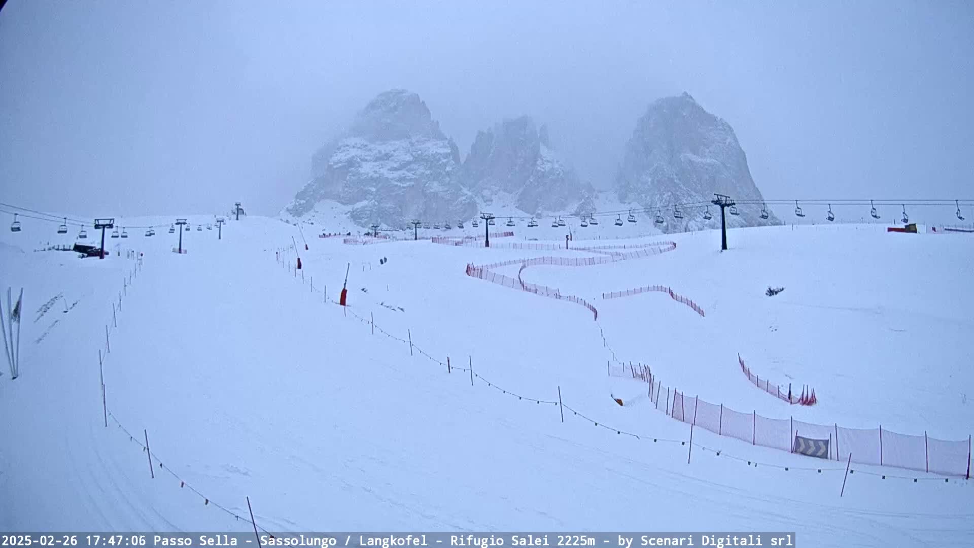A snowy ski slope with chairlifts and red safety nets is visible beneath a hazy, overcast sky, with snow-covered mountains in the background.