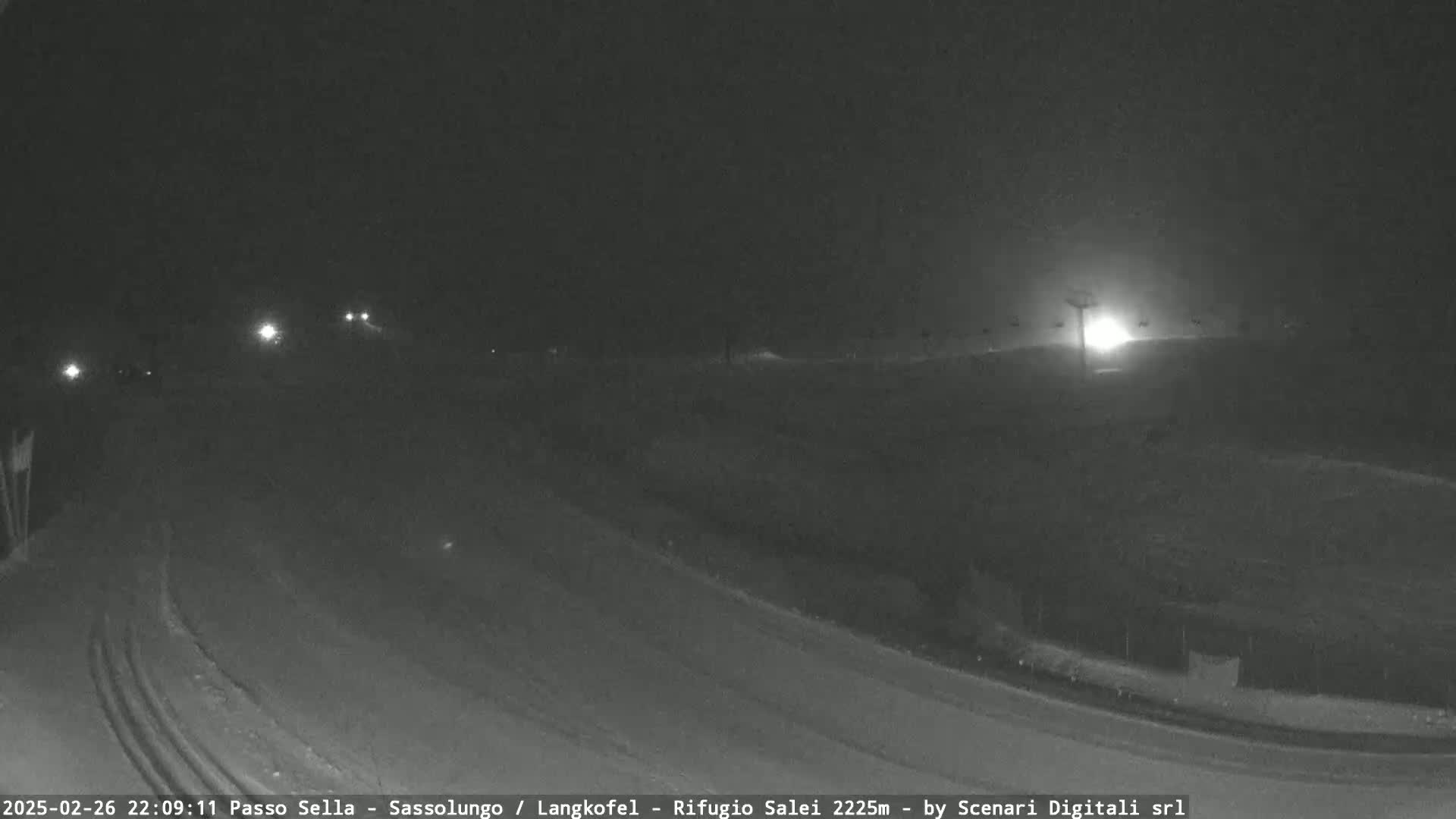A snow-covered road curves gently at night, with a few distant headlights visible, under a dark sky.