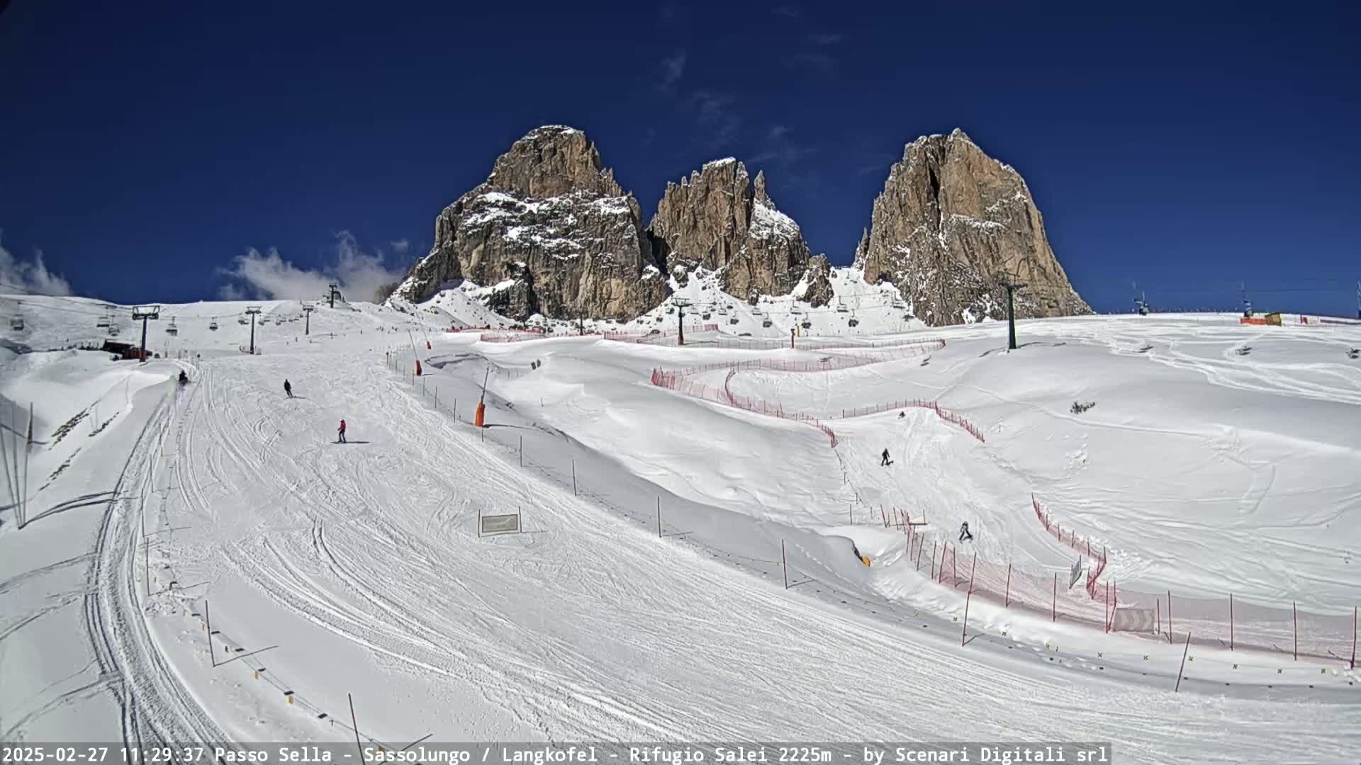 A snowy ski slope with several skiers and ski lifts, set against a backdrop of towering rocky mountains under a clear blue sky.