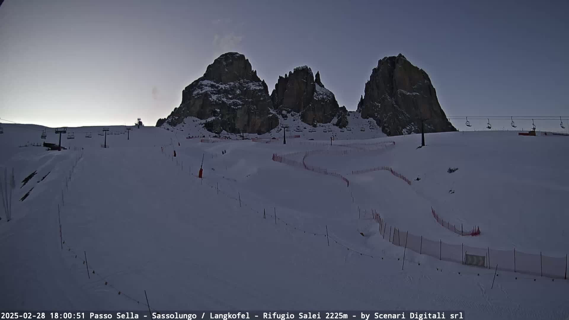 A snow-covered ski slope with chairlifts and marked runs is visible beneath a twilight sky, with imposing rocky peaks in the background.