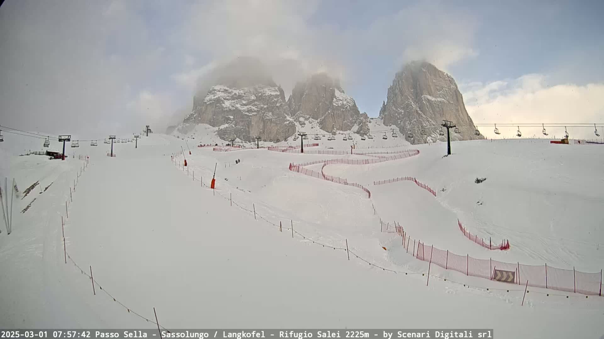 A snowy ski slope with chairlifts and red safety netting winds around snow-covered mountains partially obscured by clouds.