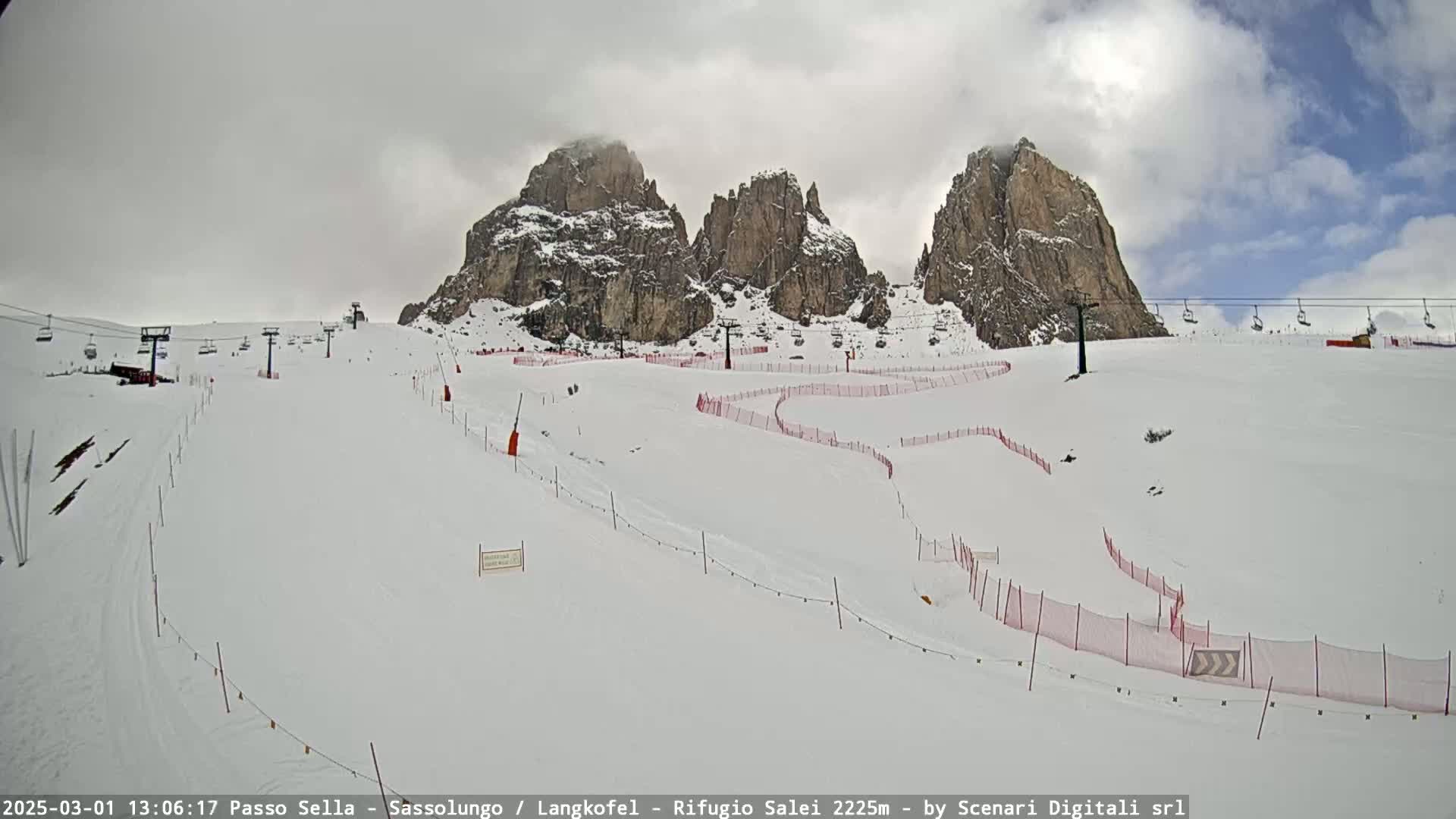 A snow-covered ski slope with several chairlifts and red safety nets winds around rocky, snow-dusted mountains under a partly cloudy sky.