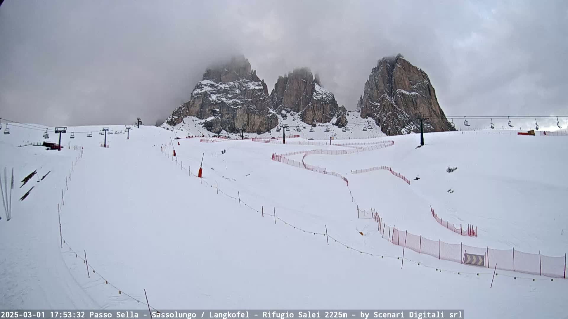 A snow-covered ski slope with chairlifts and red safety nets, set against a backdrop of rocky mountains under a cloudy sky.