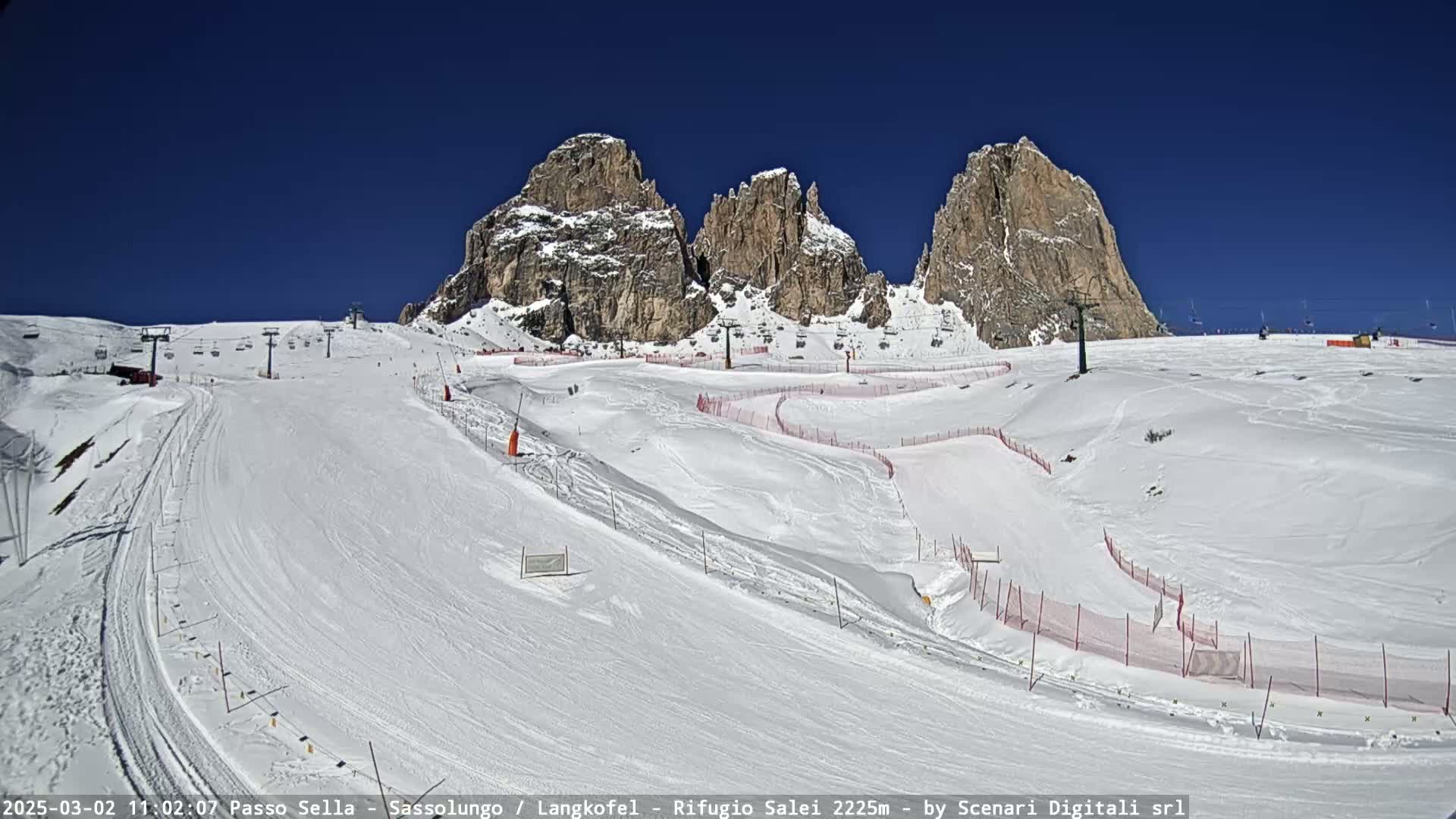 A snow-covered ski slope with ski lifts and several rocky peaks in the background under a clear blue sky.