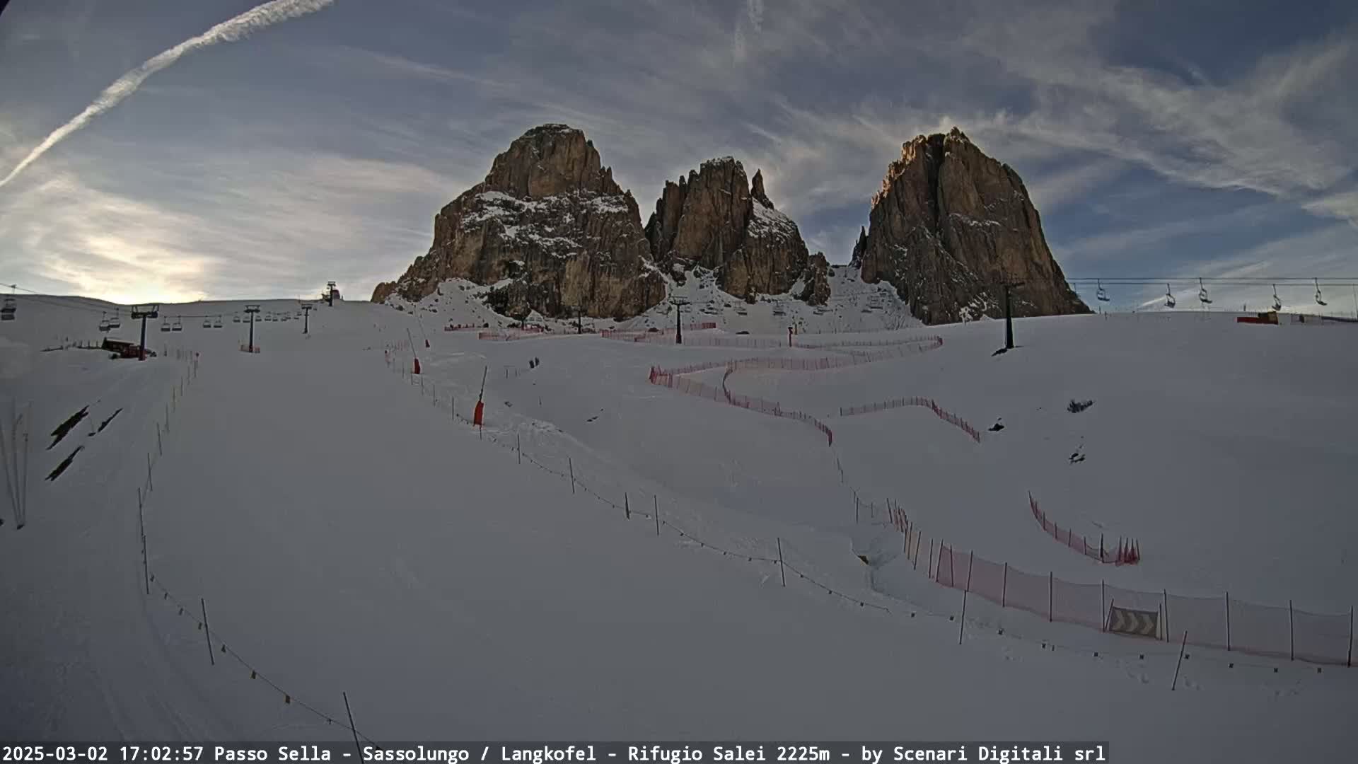 A snow-covered ski slope with chairlifts and red safety netting in front of a range of rocky mountains under a partly cloudy sky.