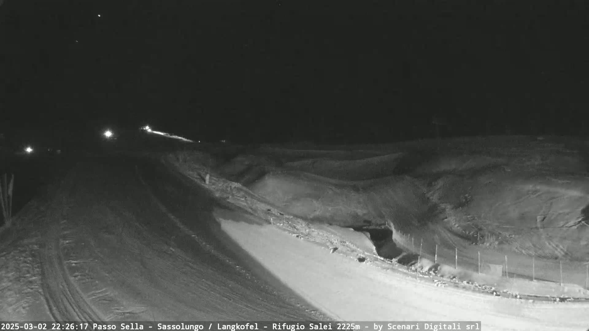 A nighttime view of a snow-covered road with a few vehicles driving on it, leading to a snow-covered landscape under a clear, dark sky.
