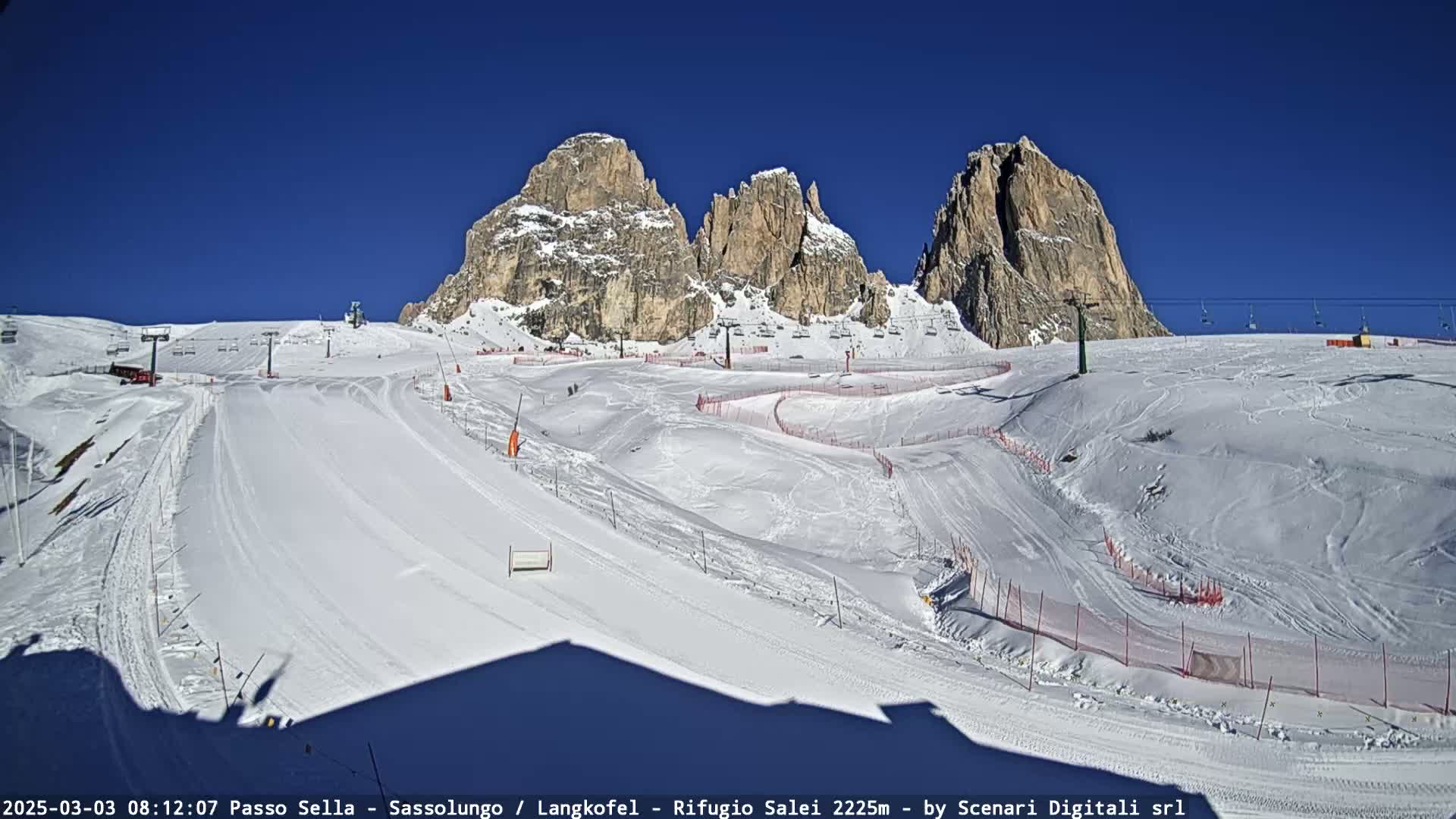A snow-covered ski slope with several ski lifts and mountain peaks in the background under a clear blue sky.