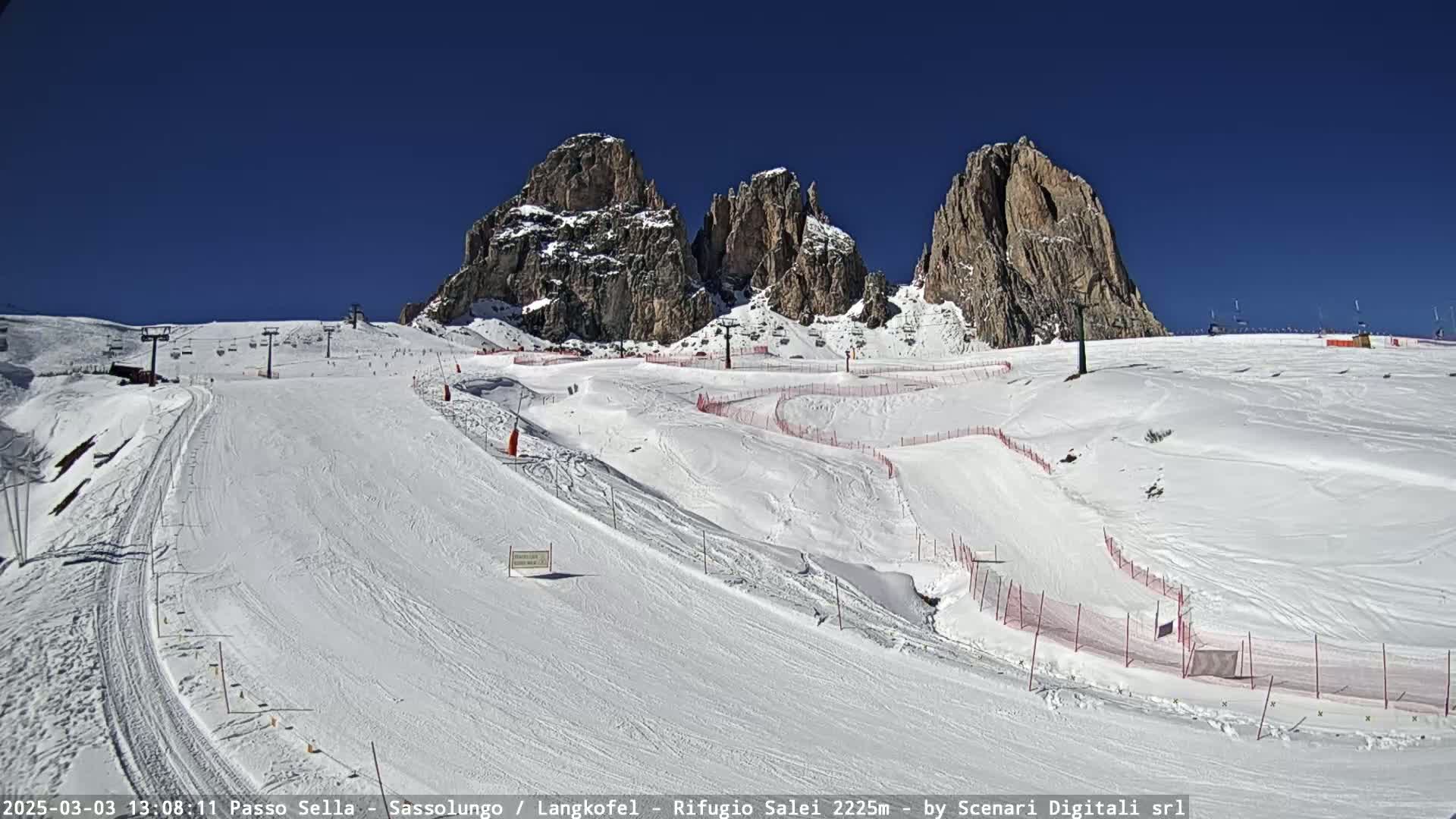 A snow-covered ski slope winds beneath towering, snow-dusted rock formations on a clear, sunny day.