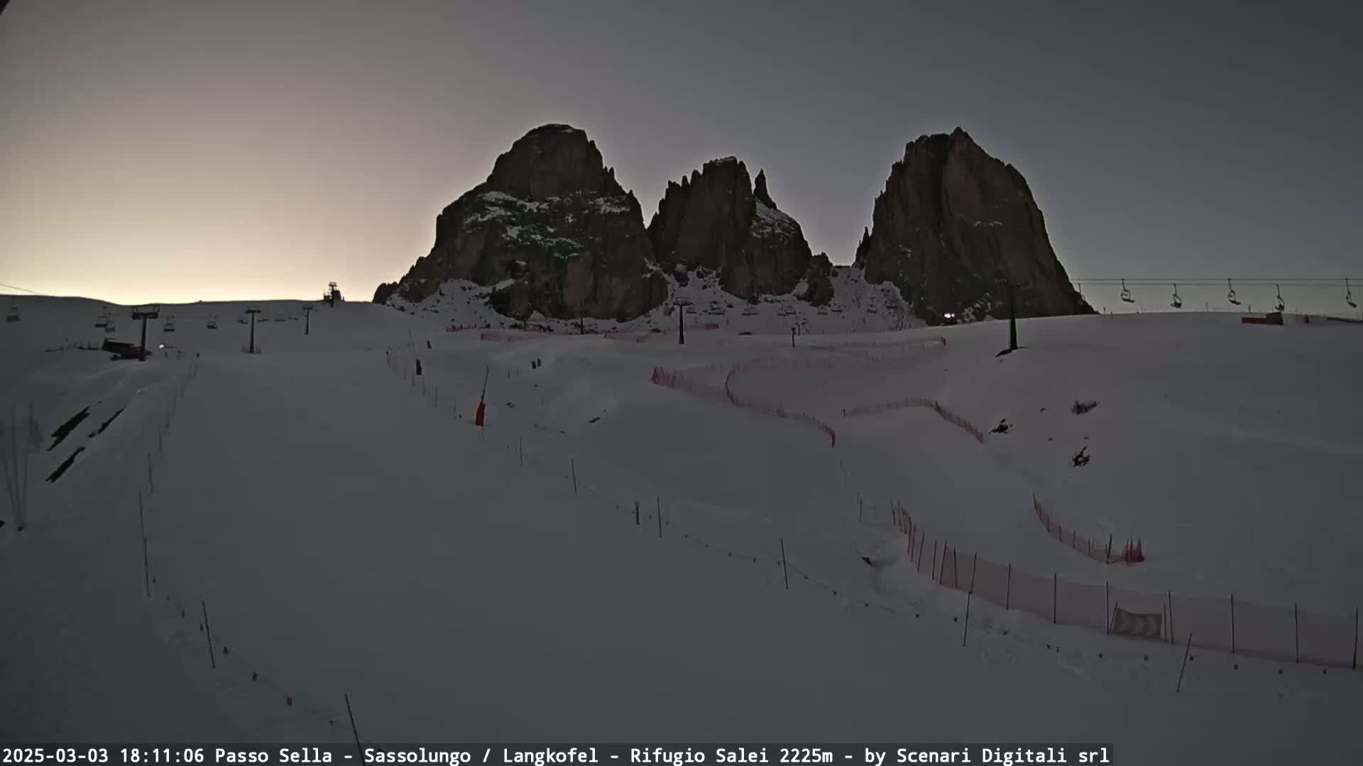 A snow-covered ski slope with ski lifts and fences is situated beneath a group of dark, rocky mountains under a dusky sky.