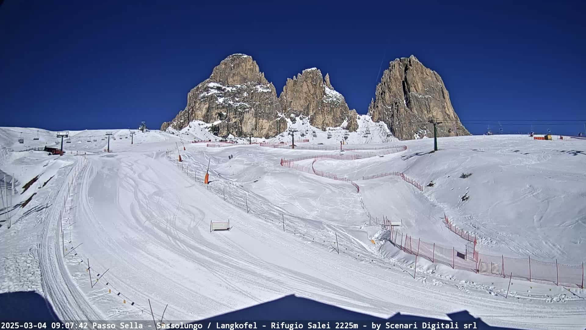 A snow-covered ski slope with ski lifts and several tall, rocky mountains in the background under a clear blue sky.