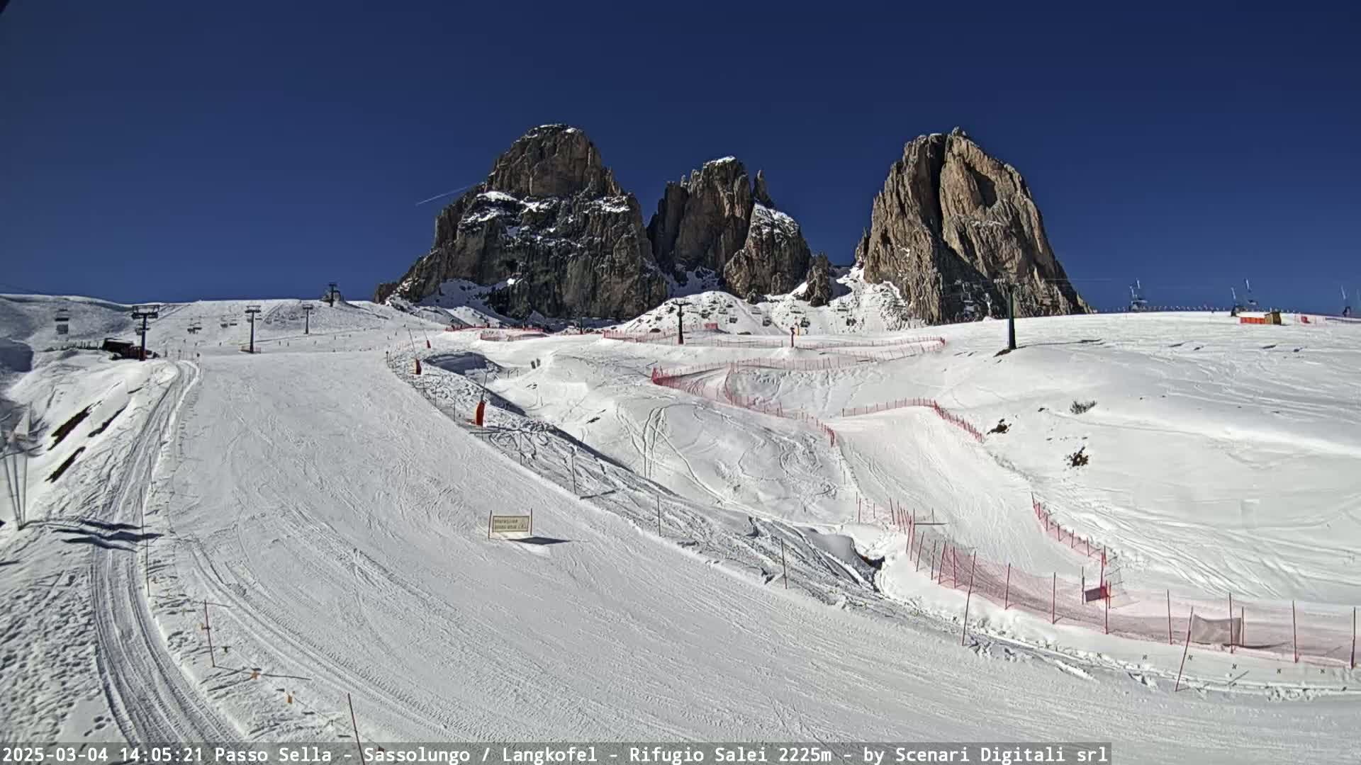 A snow-covered ski slope with ski lifts and steep, rocky mountains in the background under a clear blue sky.
