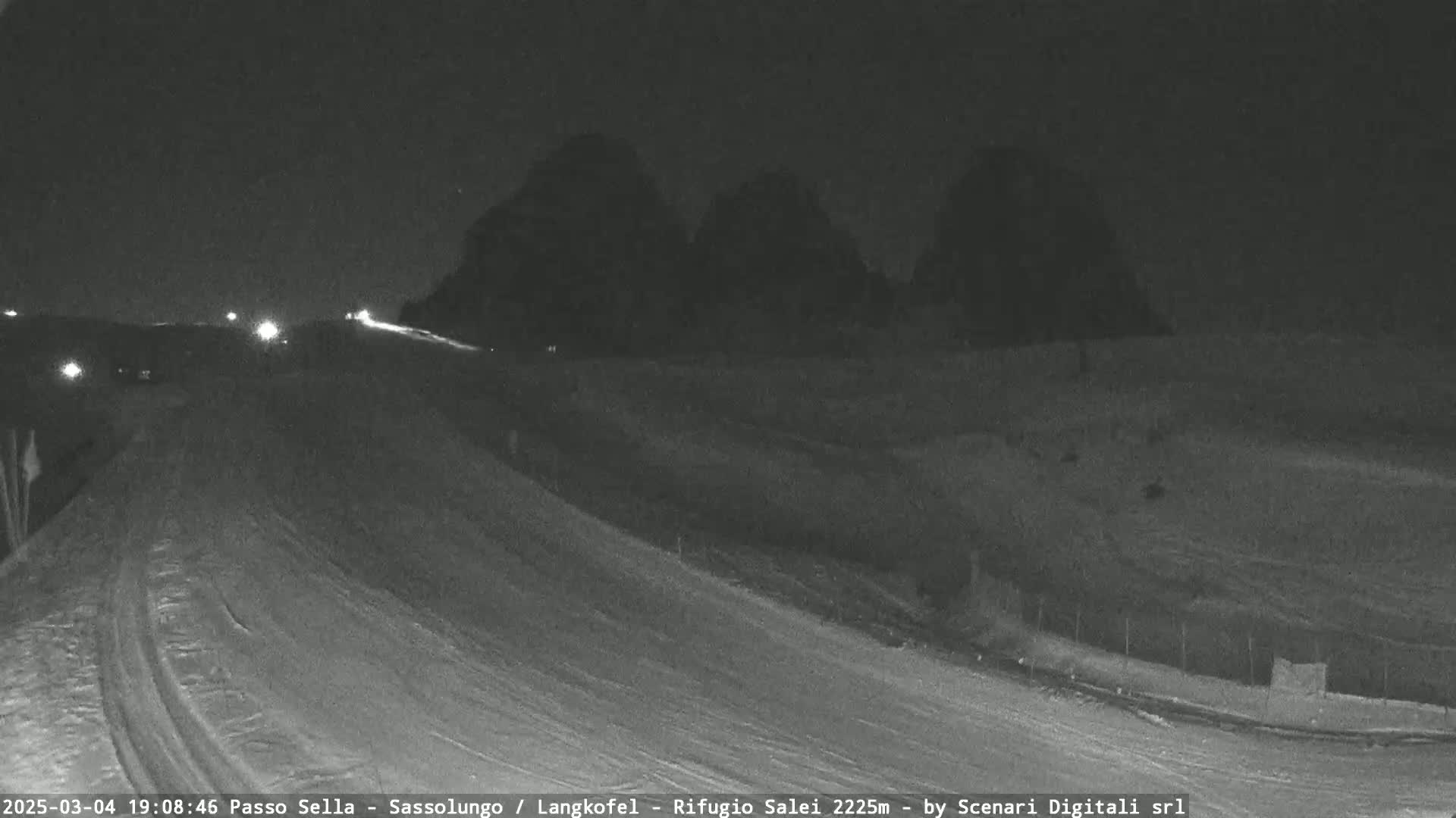 A snow-covered road at night with a few vehicles driving on it, and dark silhouettes of trees and hills in the background under a dark sky.
