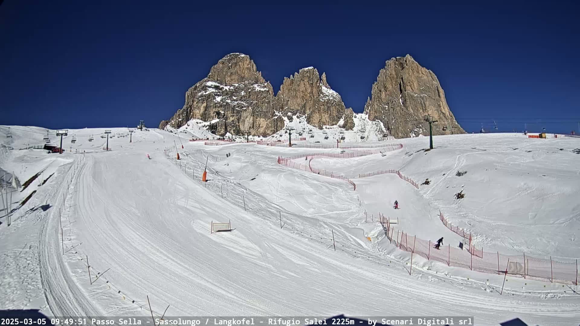 A sunny day at a snow-covered ski resort shows several skiers on marked slopes beneath towering, rocky mountains.