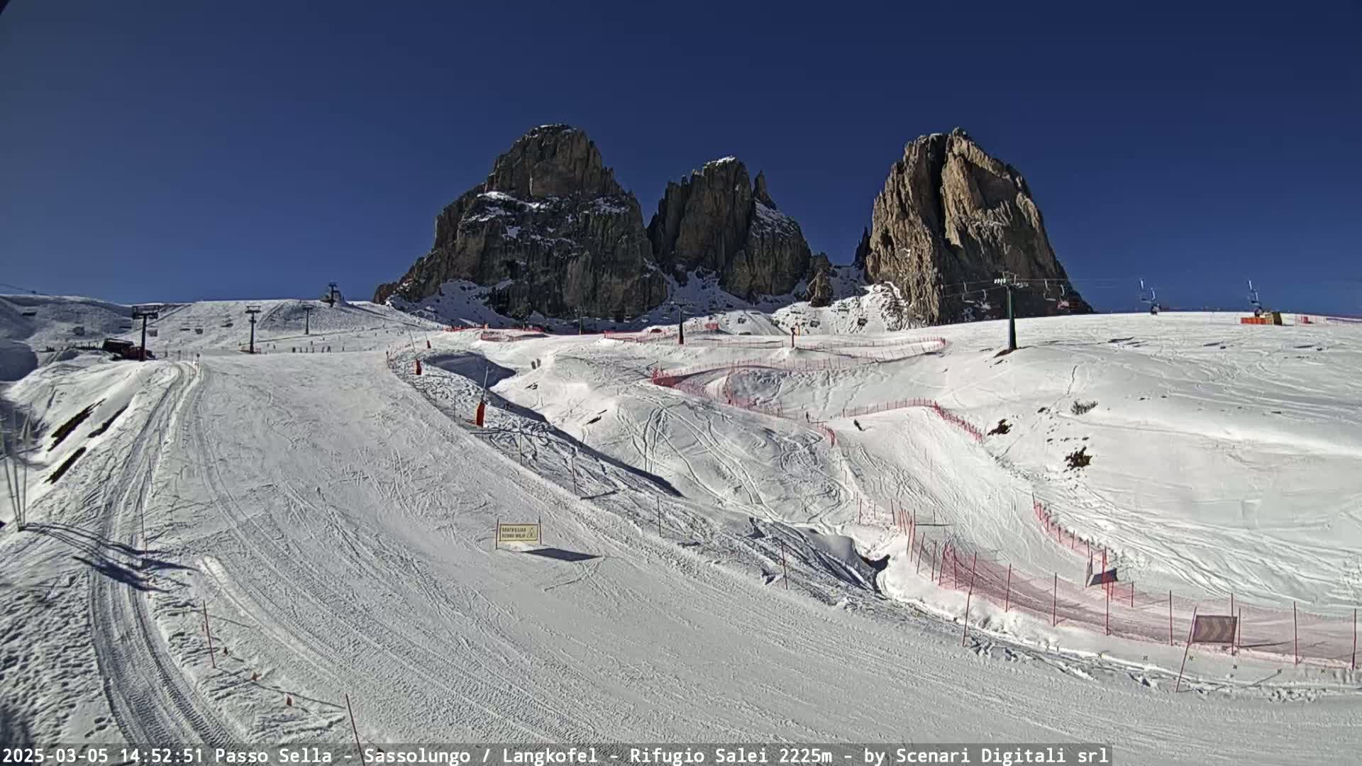 A sunny, snow-covered ski slope with marked trails winds around rocky mountain peaks under a clear blue sky.
