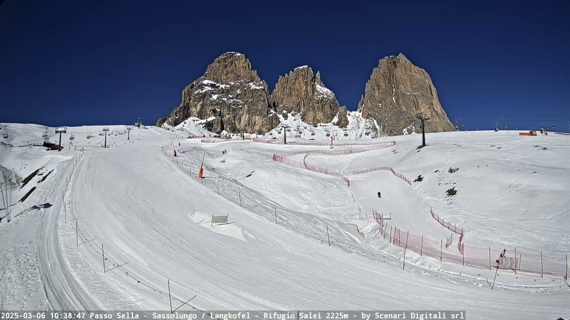 A snow-covered ski slope with several skiers visible, surrounded by rocky mountains under a clear blue sky.