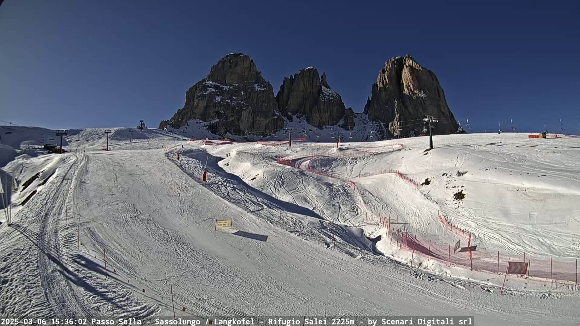 A snow-covered ski slope with several ski lifts and marked runs is situated below a backdrop of tall, rocky mountains under a clear, sunny sky.