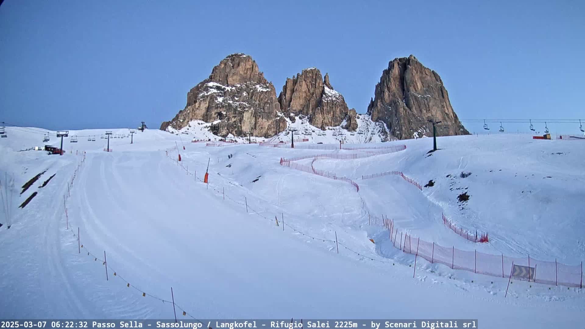 A snow-covered ski slope with several ski lifts and rocky mountains in the background under a clear blue sky.
