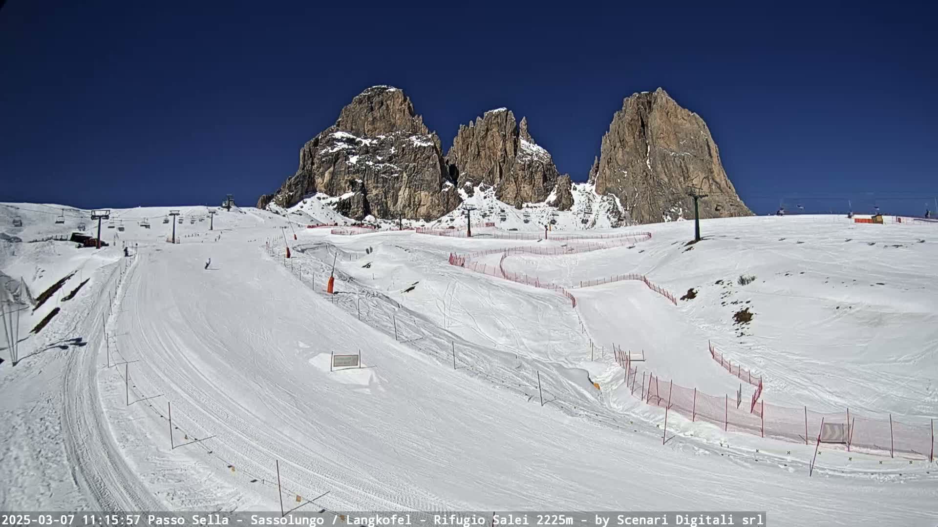A snow-covered ski slope with several ski lifts and a mountain range in the background under a clear blue sky.