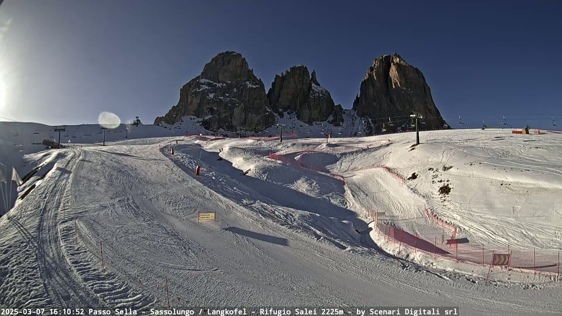 A sunny, snow-covered ski slope with several runs and ski lifts, nestled between tall, rocky mountains.