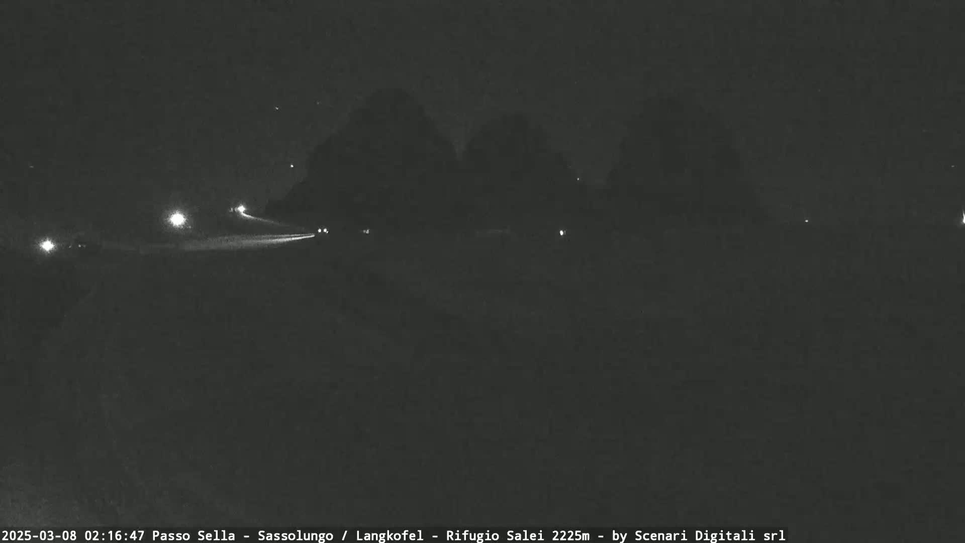 At night, a road with a few vehicles is visible, with dark, indistinct shapes of trees or hills in the background under a clear, starry sky.