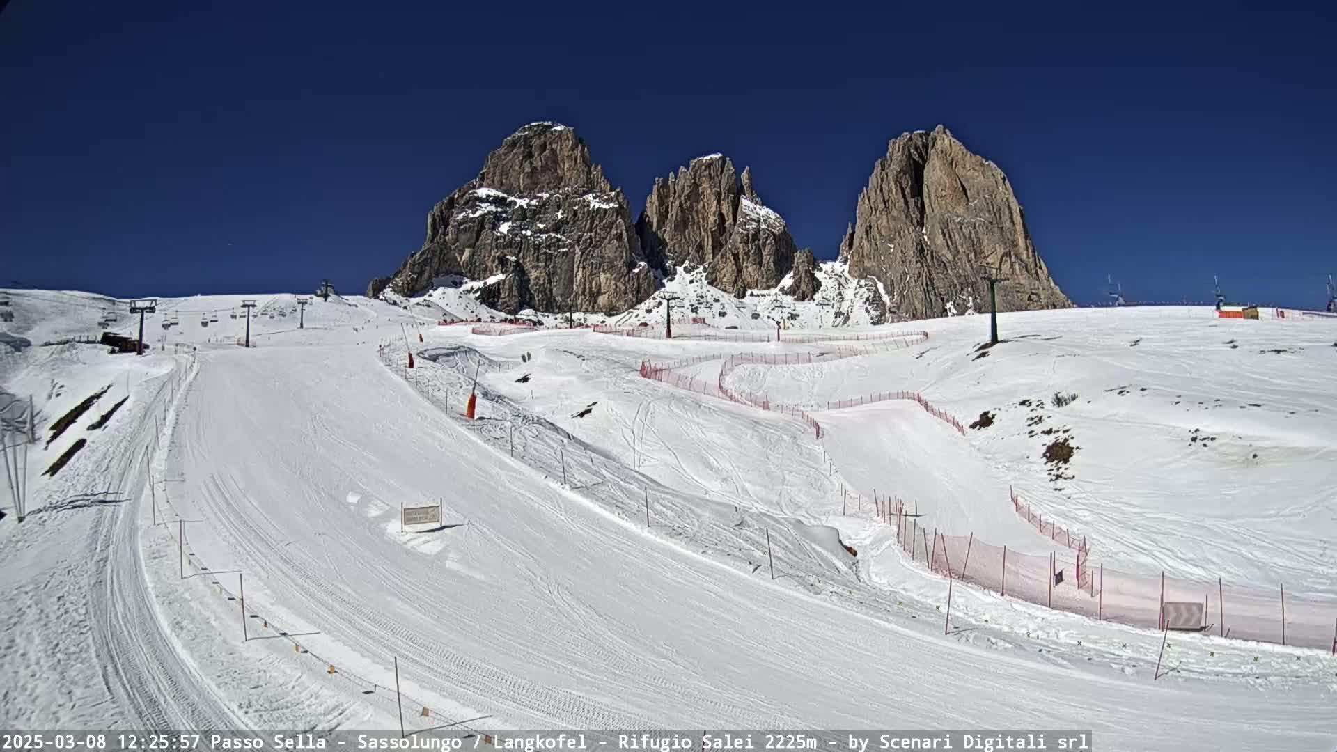 A snow-covered ski slope with several ski runs and lifts is set against a backdrop of towering, rocky mountains under a clear blue sky.