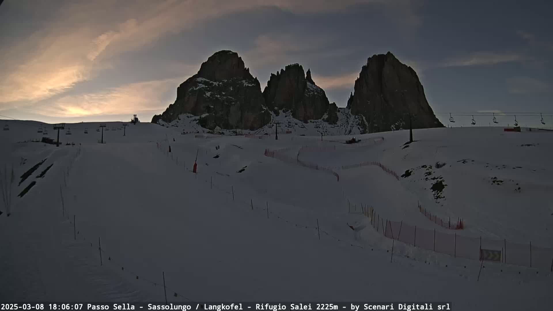 A snow-covered ski slope at dusk, with three prominent rock formations in the background and several ski lifts visible.