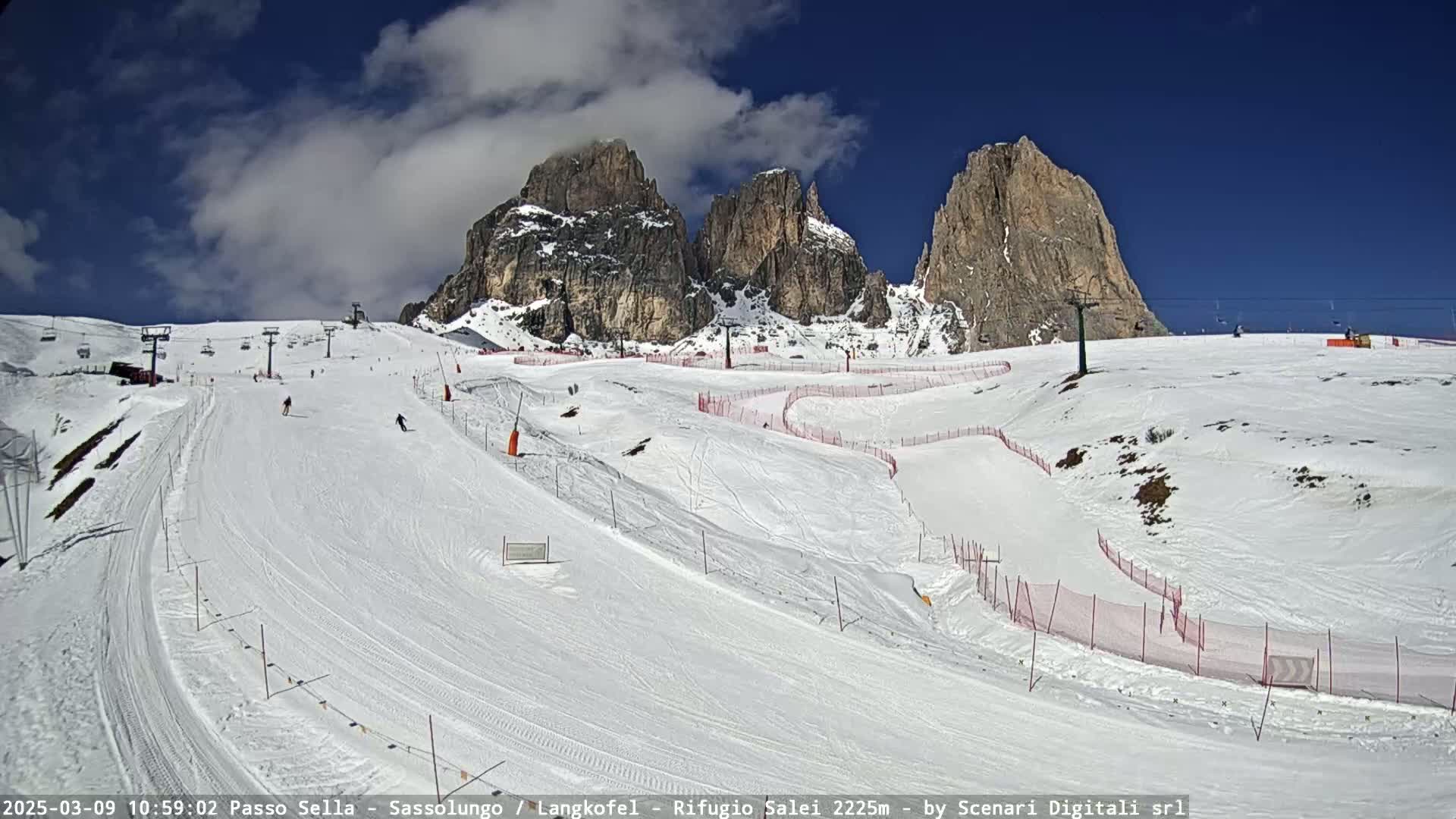 A sunny day shows a snow-covered ski slope with several skiers, ski lifts, and rocky mountains in the background.