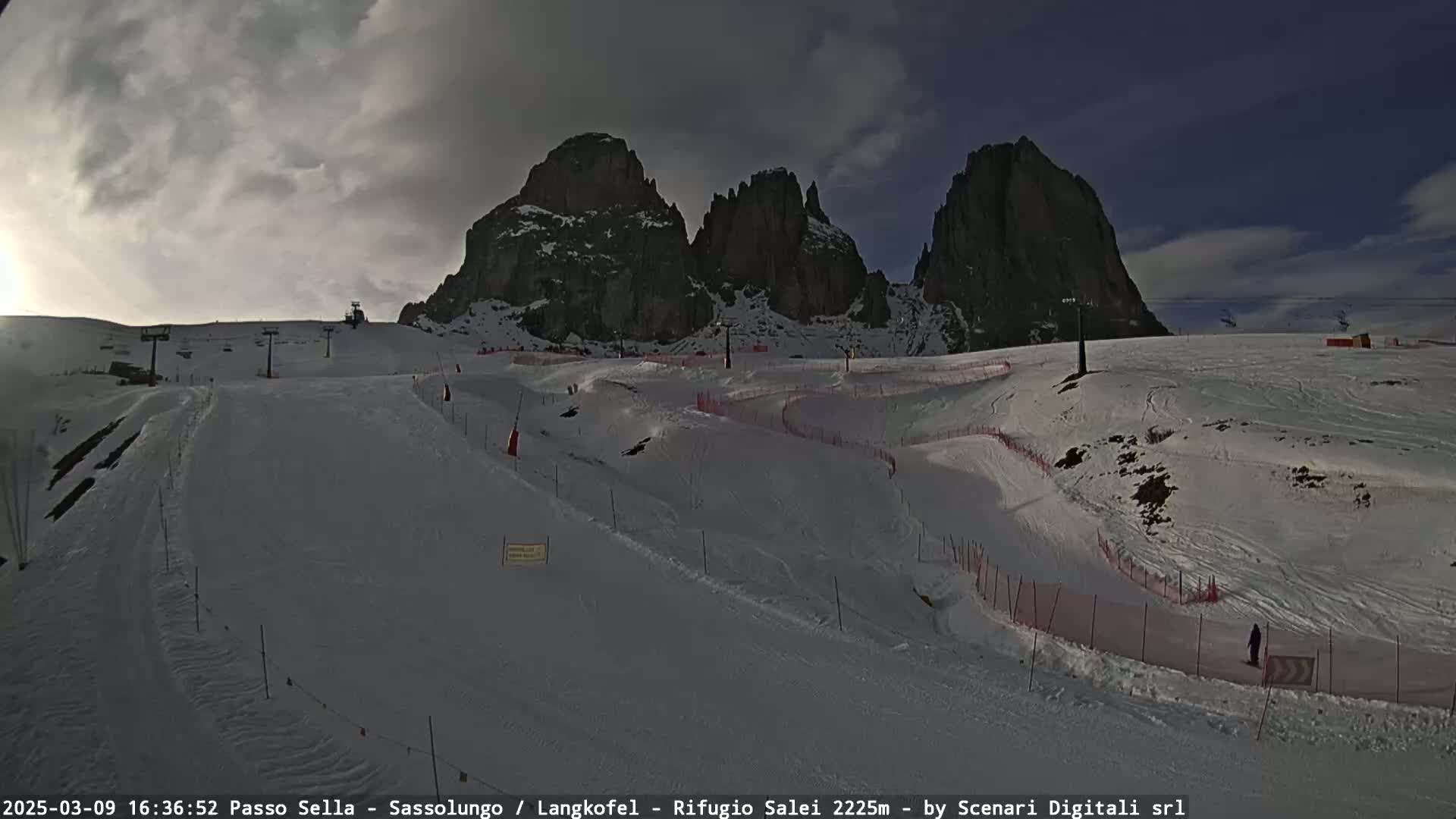 A snow-covered ski slope with winding paths and ski lifts is situated beneath a cloudy sky and imposing rocky mountains.