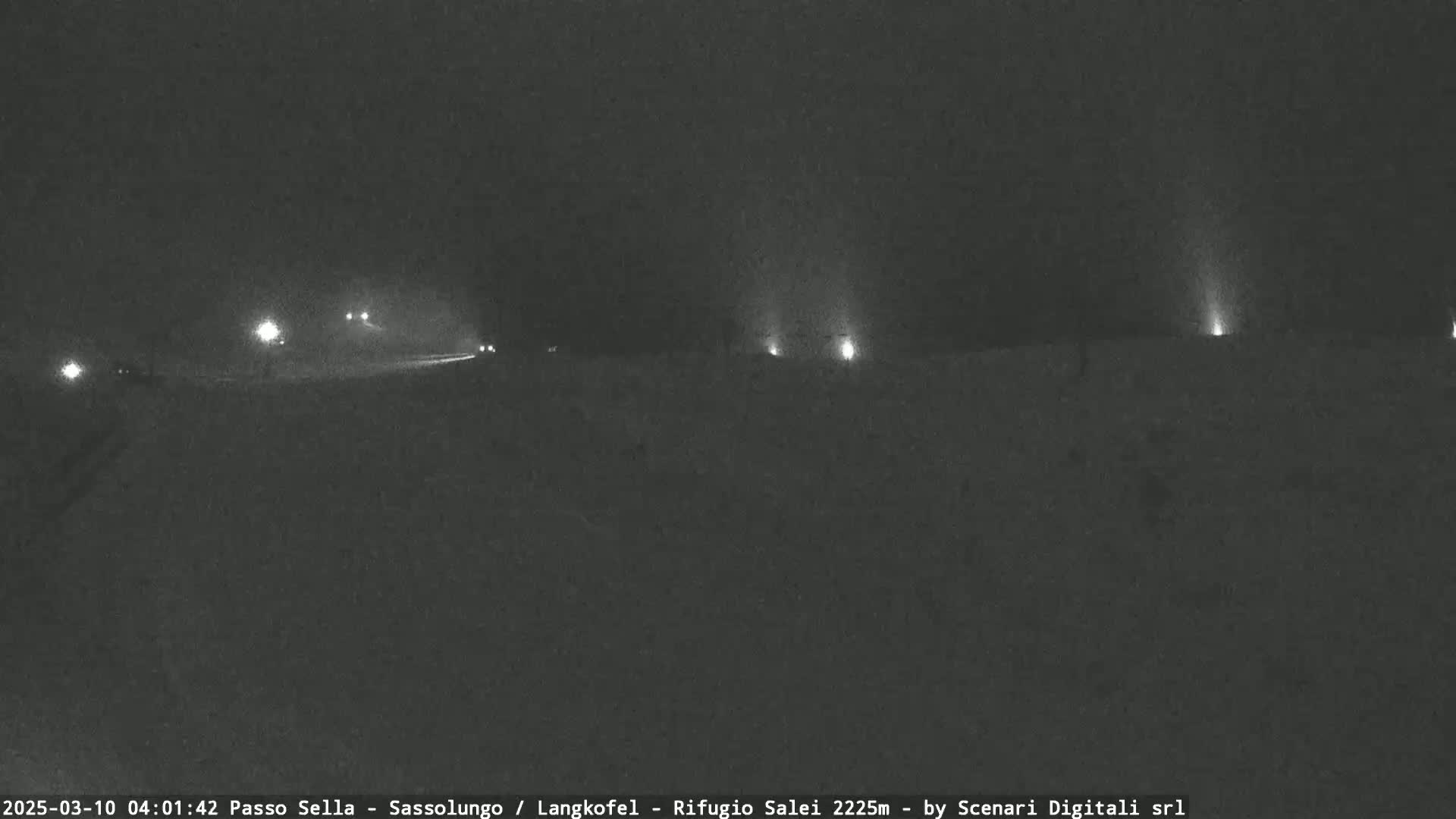 A dark, snowy road at night is dimly lit by several distant lights, with a few vehicles visible in the distance.