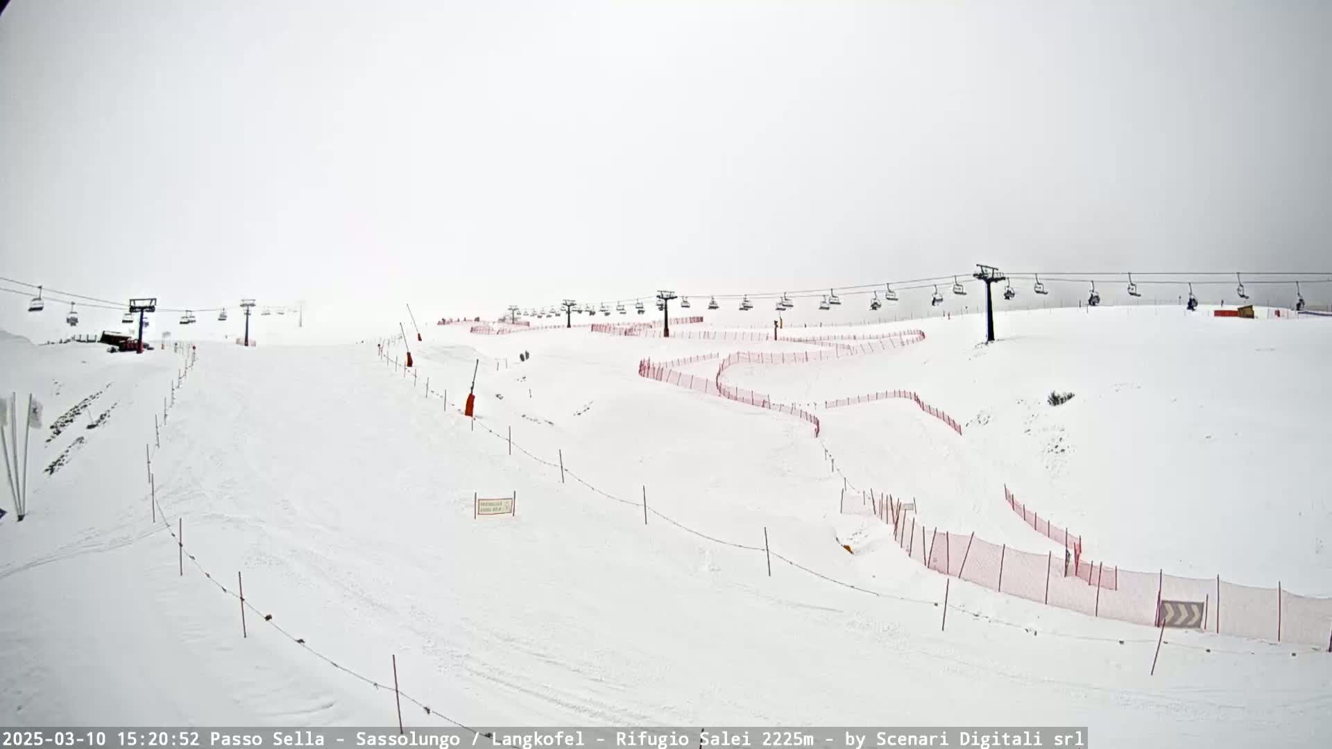 A snow-covered ski slope with chairlifts and red safety netting under an overcast sky.