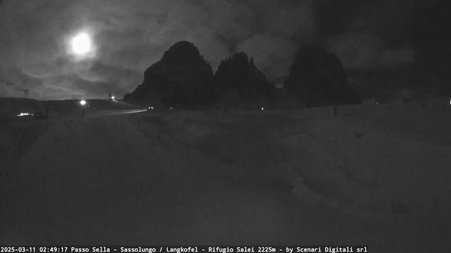 A bright moon illuminates a snow-covered landscape at night, with dark, mountainous silhouettes in the background and a few vehicles visible on a road in the foreground.