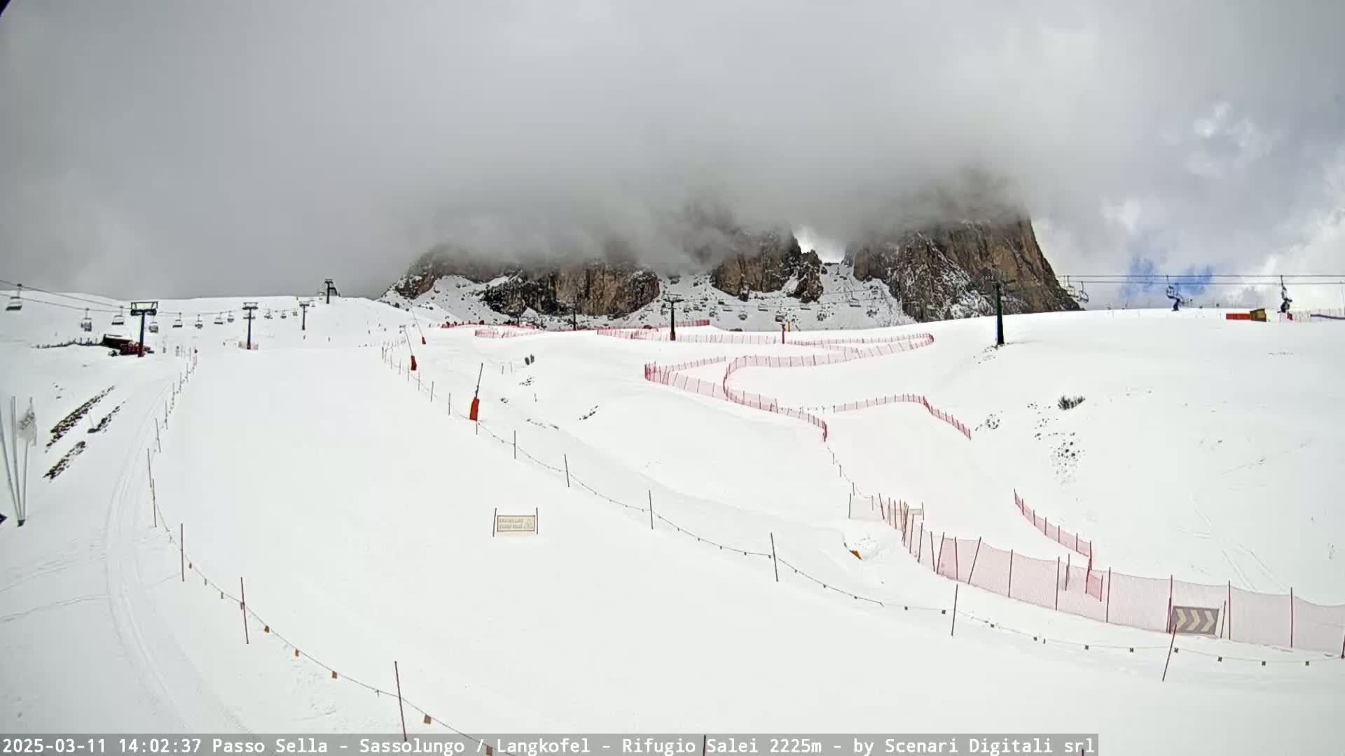 A snow-covered ski slope with chairlifts and red safety nets is visible beneath a cloudy sky.