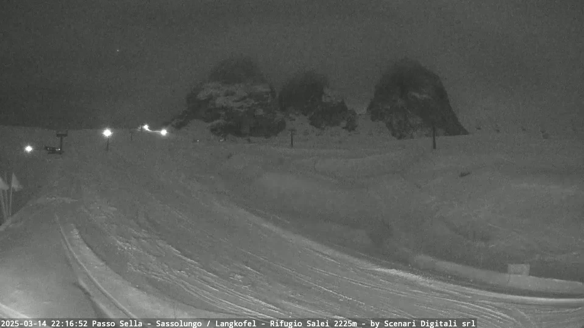 A nighttime, snowy scene shows a gently sloping snow-covered landscape with two rocky mountain peaks in the background under a dark, overcast sky.