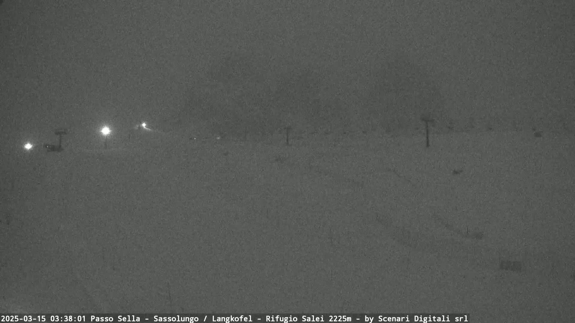 A nighttime, snowy scene shows a dimly lit roadway with several lights visible in the distance.