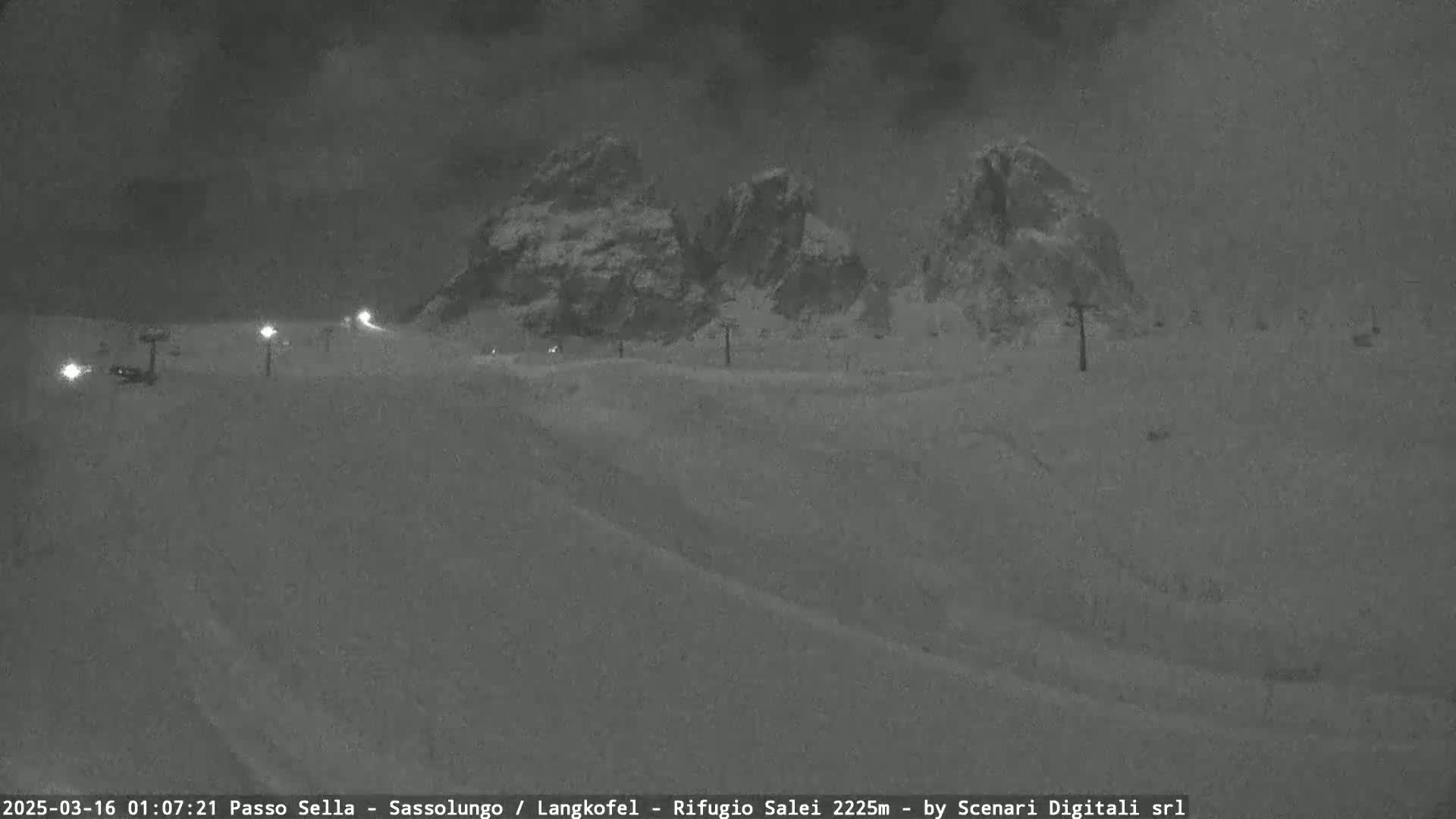 A nighttime, snow-covered landscape shows a mountain range under a dark, cloudy sky, with faintly lit ski lifts in the foreground.