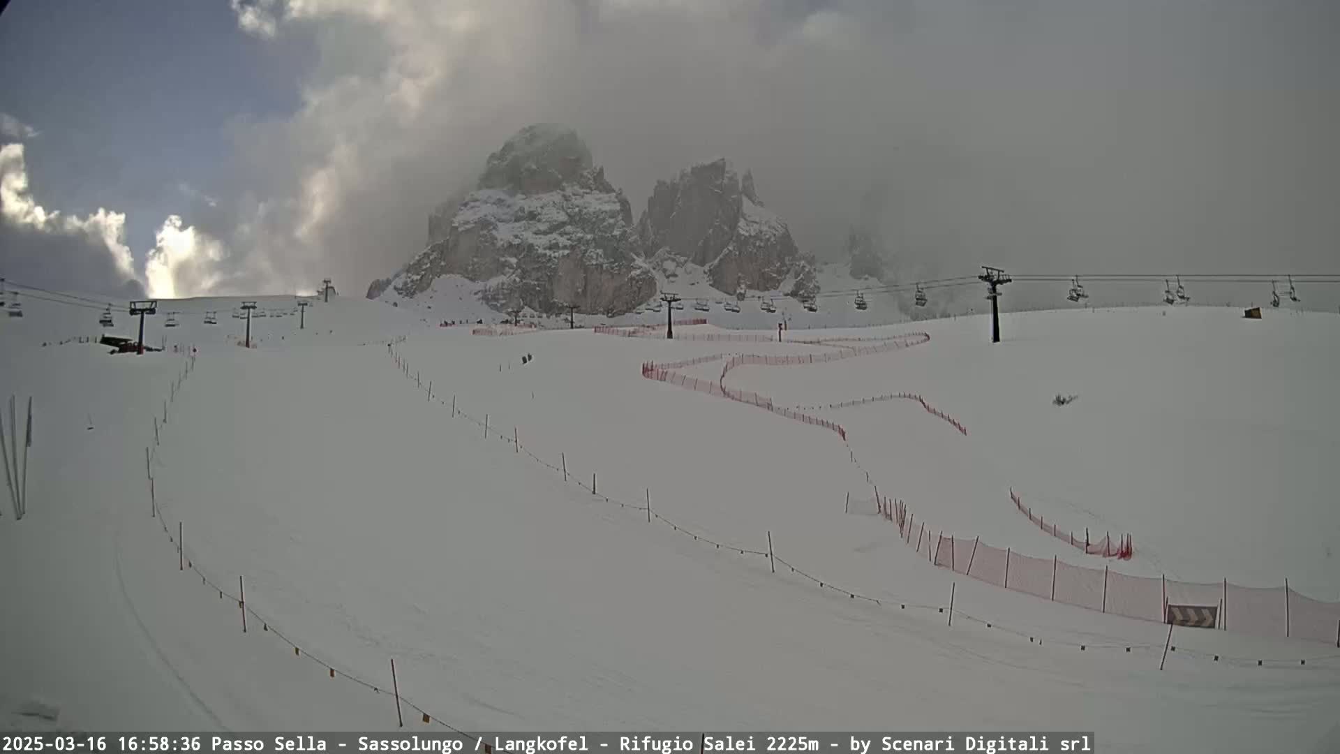 A snow-covered ski slope with chairlifts and red safety netting stretches towards rocky, snow-dusted mountains under a cloudy sky.