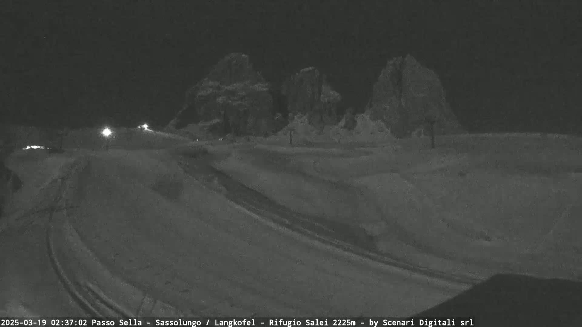 A nighttime, snowy scene shows a road winding through a mountainous area under a dark sky.