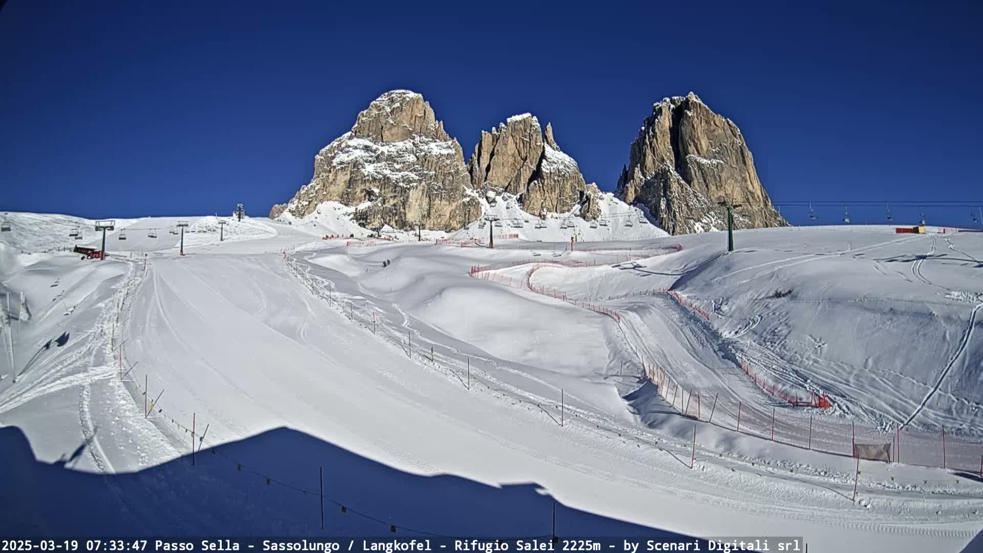 A snow-covered ski slope with marked runs and chairlifts is situated beneath three towering, rocky mountain peaks under a clear, bright blue sky.