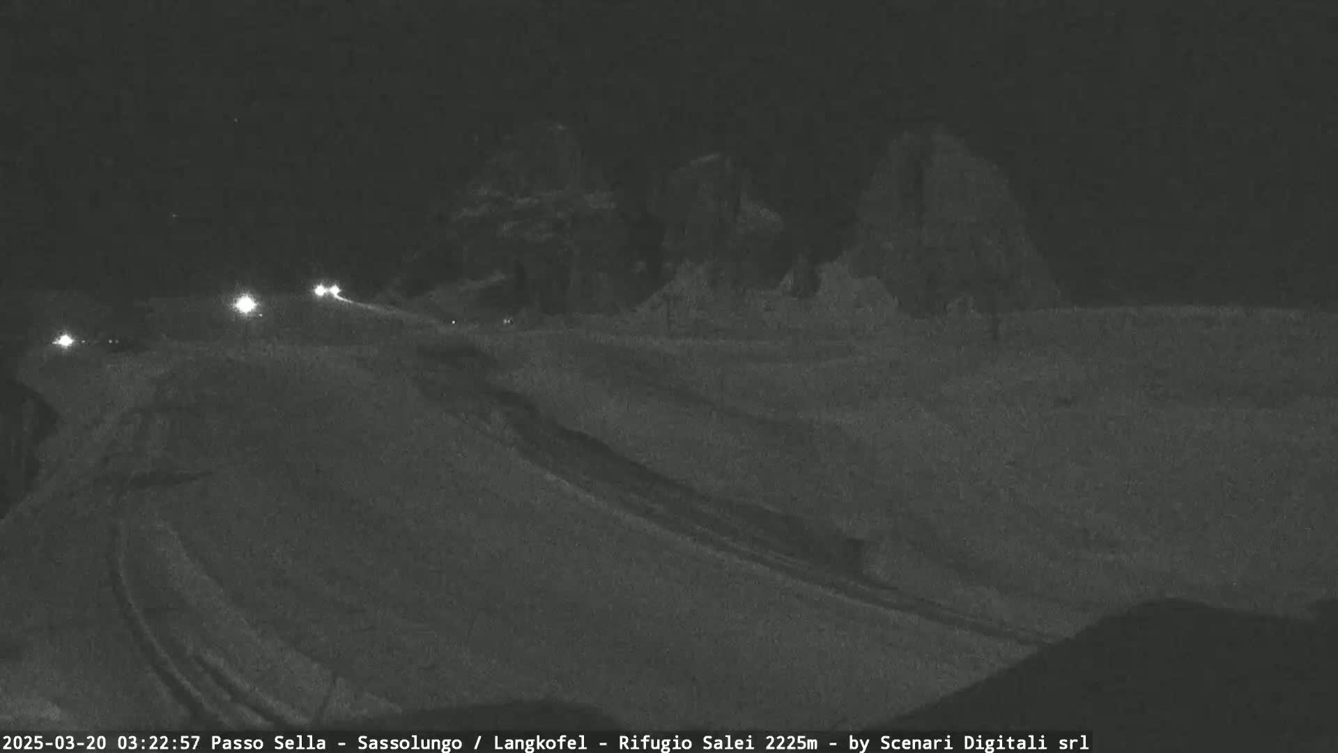 A nighttime, snowy road curves gently to the right, with a few vehicle headlights visible in the distance and dark, snow-covered hills in the background.